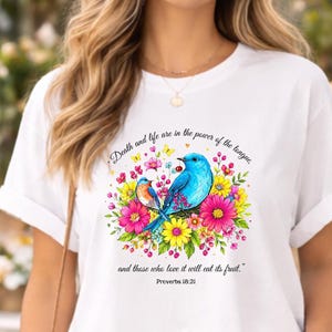 Puede incluir: Camiseta blanca con una ilustración colorida de pájaros y flores. El diseño incluye el texto "Death and life are in the power of the tongue... Proverbios 18:21."