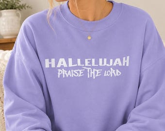 May include: A light purple crewneck sweatshirt featuring the white text HALLELUJAH PRAISE THE LORD printed across the chest. The garment has long sleeves and a ribbed collar, showcasing a simple design with bold lettering on the front.