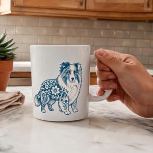 May include: White ceramic mug featuring a blue and white illustration of a dog with floral patterns. The dog's fur is detailed, and the mug has a curved handle. The mug is held by a person's hand.