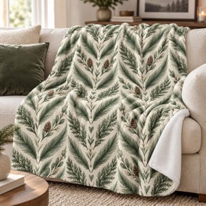 May include: A cream-colored throw blanket featuring a repeating pattern of green pine branches, brown pinecones, and small white berries. The blanket has a soft, white, textured underside visible at the folded corner, draped over a sofa.