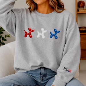 May include: A heather gray crewneck sweatshirt featuring three balloon animal dogs in red, white, and blue across the chest. The left sleeve displays the text 250 next to a small American flag graphic. The garment has long sleeves and a ribbed neckline.