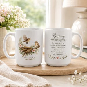 May include: Two white ceramic mugs featuring a bird nest illustration with two brown birds, pink flowers, and green leaves. One mug displays the text "Be strong and courageous" followed by the full verse from Deuteronomy 31:6 in black script.