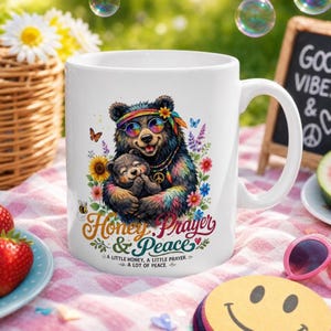 May include: White ceramic mug featuring a colorful illustration of a bear and cub embracing, wearing sunglasses and a headband. The mug has the words "Honey, Prayer & Peace" and the phrase "A little honey, a little prayer, a lot of peace."