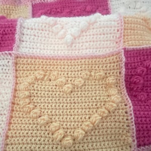 Puede incluir: Una manta de ganchillo hecha a mano con un diseño de patchwork. La manta presenta cuadrados en tonos de rosa, melocotón y blanco. Algunos cuadrados tienen patrones de corazón y flores en relieve. Los bordes están acabados con un ribete festoneado.