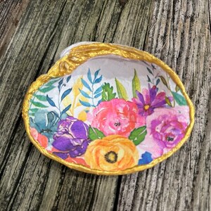 May include: A white seashell with a gold rim and a floral pattern. The shell is decorated with pink, purple, yellow, and green flowers.