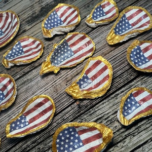 May include: A collection of oyster shells painted with the American flag design. The shells are gold-rimmed and arranged on a wooden surface.