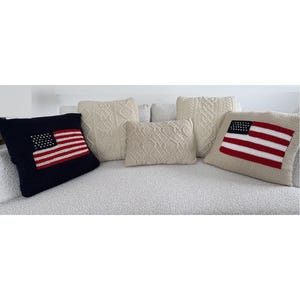 May include: A collection of five decorative throw pillows. Two pillows feature a knitted American flag design, one on a navy blue background and one on a beige background. Three pillows feature an off-white cable knit pattern in square and rectangular shapes.