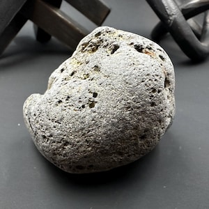 Könnte beinhalten: Ein grauer, poröser Stein mit rauer Textur. Der Stein ist unregelmäßig geformt und scheint ein natürliches Objekt zu sein. Andere dunkle Objekte befinden sich im Hintergrund.