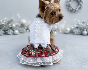 May include: A small dog wearing a white sleeveless top with three buttons and a white bow. The skirt features a red, green, and gold plaid pattern with snowflake designs, layered over a white polka-dot tulle ruffle with a metal D-ring attached.