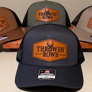 Puede incluir: Cuatro gorras de béisbol de diferentes colores con un parche de cuero marrón que dice "Throwin' Rows" con un logotipo de cabeza de ciervo. Las gorras son marrón, verde, gris y negra.
