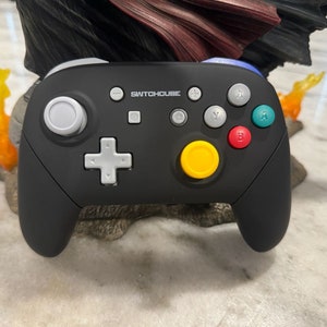 May include: A black Switchouse brand video game controller with a yellow button, gray buttons, and blue and green buttons. The controller is sitting on a brown surface.