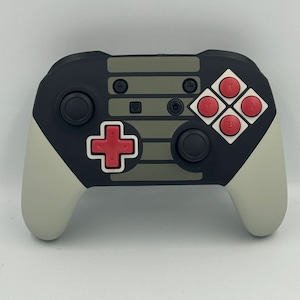 May include: A black and gray Nintendo Switch controller with a custom design featuring a gray and black striped pattern reminiscent of the classic Nintendo Entertainment System. The controller has red buttons and a red plus button.