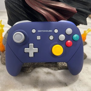 May include: A purple and grey Nintendo Switch controller with the text "SWITCHCUBE" printed on the front. The controller has a yellow button, a blue button and a green button.