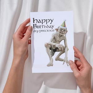 May include: A birthday card with a Gollum illustration wearing a party hat. The card reads "happy Birthday my precious" in a stylized font. The card is held against a white background.