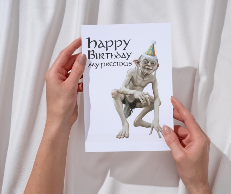 May include: A birthday card with a Gollum illustration wearing a party hat. The card reads "happy Birthday my precious" in a stylized font. The card is held against a white background.