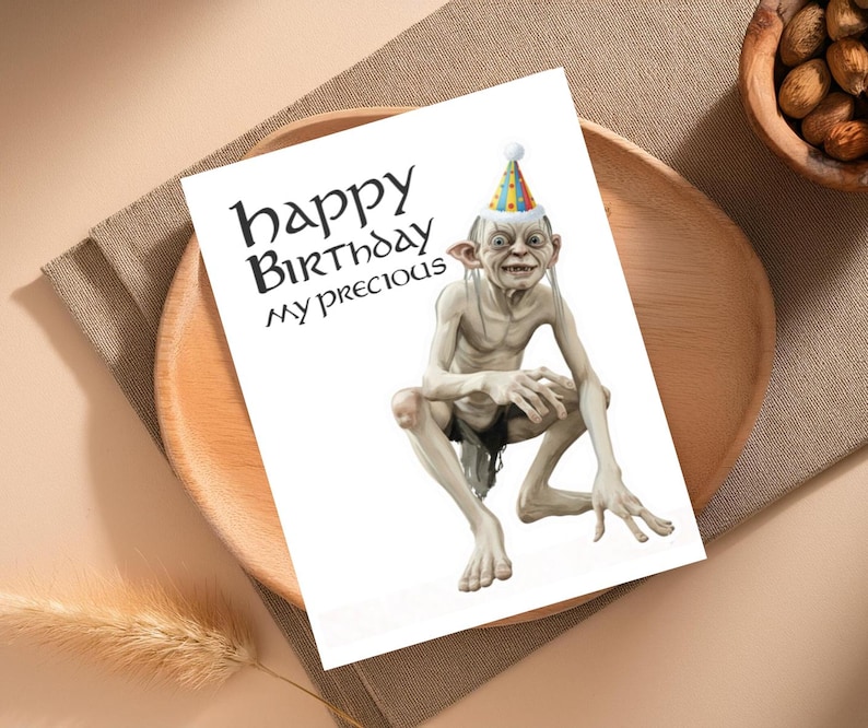May include: A white birthday card with the text "happy Birthday my precious" in black. The card features a cartoon illustration of Gollum wearing a party hat. The card is on a wooden plate.