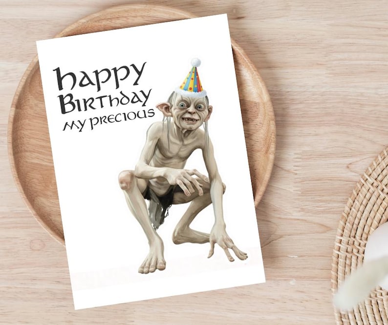 May include: A white birthday card with the words "happy Birthday my precious" in black. The card shows an illustration of Gollum from The Lord of the Rings wearing a party hat. The card is on a wooden plate.