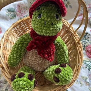 May include: A green plush turtle wearing a red hat and scarf, in a woven basket. The turtle has brown accents on its feet and a tan belly. The plush toy is made of a soft, textured material.