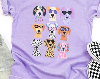 May include: A violet t-shirt featuring a graphic print of nine dogs wearing colorful sunglasses, accompanied by a pair of light blue denim shorts with frayed hems and a pair of black and white checkered slip-on sneakers. The text violet is visible.