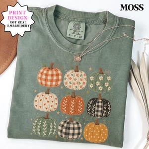 May include: A moss green crew neck t-shirt featuring a graphic print of nine pumpkins in various patterns including gingham, floral, heart, and polka dot designs. The shirt has a Comfort Colors label and the text MOSS and PRINT DESIGN NOT REAL EMBROIDERY.