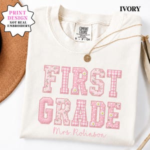 May include: An ivory t-shirt featuring a printed design of the text FIRST GRADE in pink gingham, floral, and polka dot patterns with small daisy accents. Below the text, the name Mrs. Robinson is printed in a cursive pink script font.