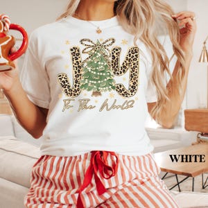 May include: White t-shirt with a Christmas-themed graphic. The design features the word "JOY" in a leopard print font, a decorated Christmas tree, and the phrase "To The World" in gold script. The shirt is paired with red and white striped pajama pants.