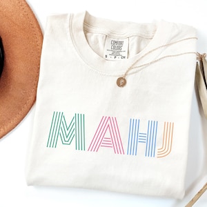 May include: Cream-colored t-shirt with the word "MAUI" in colorful striped letters. The shirt has a crew neck and short sleeves. A gold necklace with a pendant is draped on the shirt. A brown hat is partially visible.