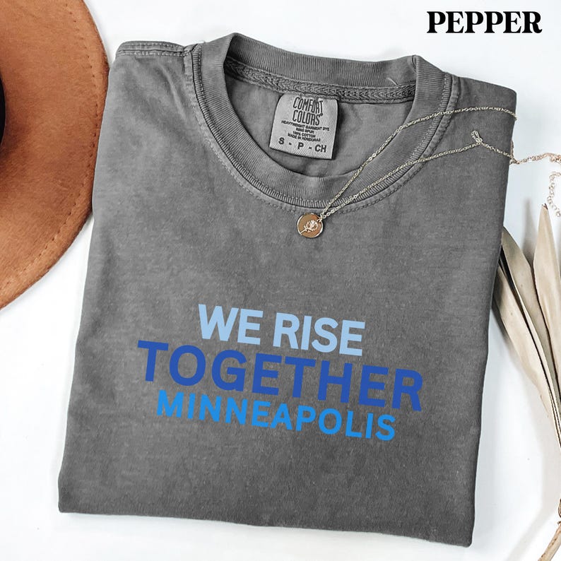 May include: A grey t-shirt with the text "WE RISE TOGETHER MINNEAPOLIS" in blue. The shirt has a crew neck and short sleeves. A silver necklace with a pendant is draped across the shirt. The shirt is folded on a white surface.