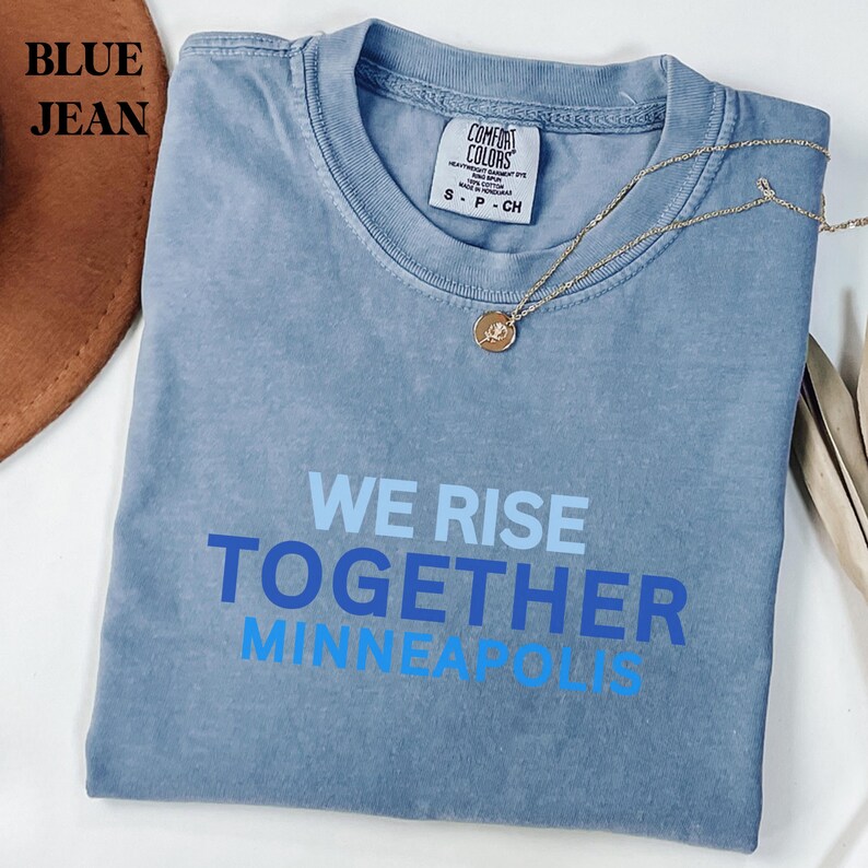 May include: A blue jean Comfort Colors t-shirt with the text "WE RISE TOGETHER MINNEAPOLIS" in blue and light blue. A gold necklace with a pendant is draped across the shirt.
