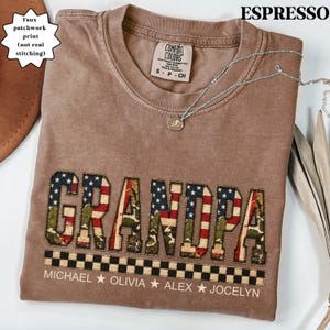 May include: Espresso-colored t-shirt with the word "GRANDPA" in a faux patchwork design featuring American flag and camouflage patterns. The shirt includes names below the word "GRANDPA". A silver necklace with a pendant rests on the shirt.
