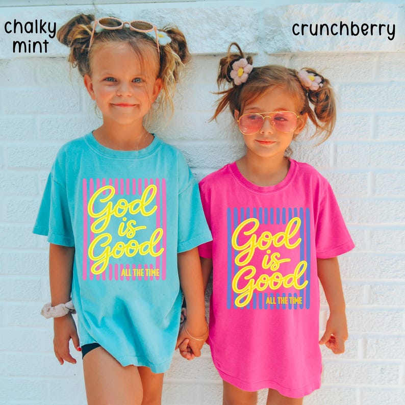 May include: Two short-sleeve t-shirts, one in chalky mint and one in crunchberry pink. Each shirt features a graphic print with vertical stripes and the yellow text God is Good All The Time. The mint shirt has pink stripes, while the pink shirt has blue stripes.