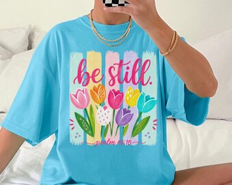Puede incluir: Una camiseta azul brillante con un gráfico de tulipanes coloridos en rosa, naranja, amarillo y morado. El texto "be still." aparece en cursiva rosa sobre las flores y "psalm 46:10" en una letra rosa más pequeña bajo el diseño floral.