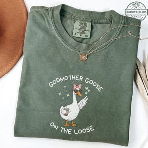 May include: A moss green Comfort Colors t-shirt featuring a graphic of a goose wearing sunglasses and a pink bow, holding a coffee cup. The text reads "Godmother Goose on the Loose". A gold necklace is draped on the shirt.