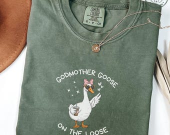 May include: A moss green Comfort Colors t-shirt featuring a graphic of a goose wearing sunglasses and a pink bow, holding a coffee cup. The text reads "Godmother Goose on the Loose". A gold necklace is draped on the shirt.