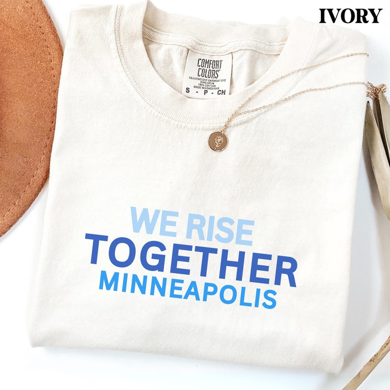 May include: An ivory-coloured t-shirt with the text "WE RISE TOGETHER MINNEAPOLIS" in shades of blue. A gold necklace with a pendant is draped across the shirt. The shirt is folded, and a brown hat is partially visible.