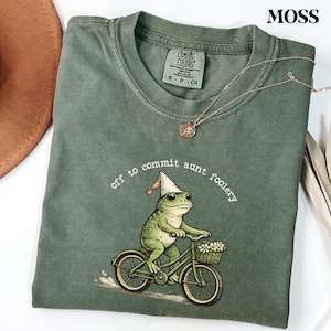 May include: Moss green t-shirt with a whimsical frog riding a bicycle, wearing a party hat, and carrying flowers in a basket. The text "off to commit aunt foolery" is printed above the frog. A silver necklace with a pendant is draped on the shirt.
