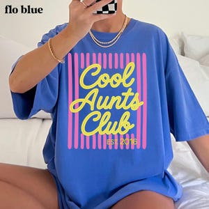 May include: A periwinkle blue t-shirt featuring a graphic design with vertical pink stripes and the yellow text "Cool Aunts Club" followed by "EST. 2016". The shirt has short sleeves and a crew neckline, showcasing a casual and relaxed fit.