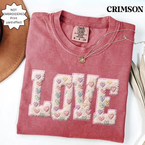 May include: Crimson-colored t-shirt with the word "LOVE" embroidered with small hearts in various colors. The shirt has a crew neck and a Comfort Colors tag. A silver necklace with a pendant is draped on the shirt.