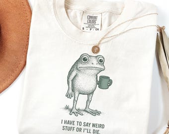 May include: Ivory-colored t-shirt with a graphic of a frog holding a mug. The frog is drawn in a detailed, vintage style. The text on the shirt reads, "I HAVE TO SAY WEIRD STUFF OR I'LL DIE." A gold necklace is draped across the shirt.