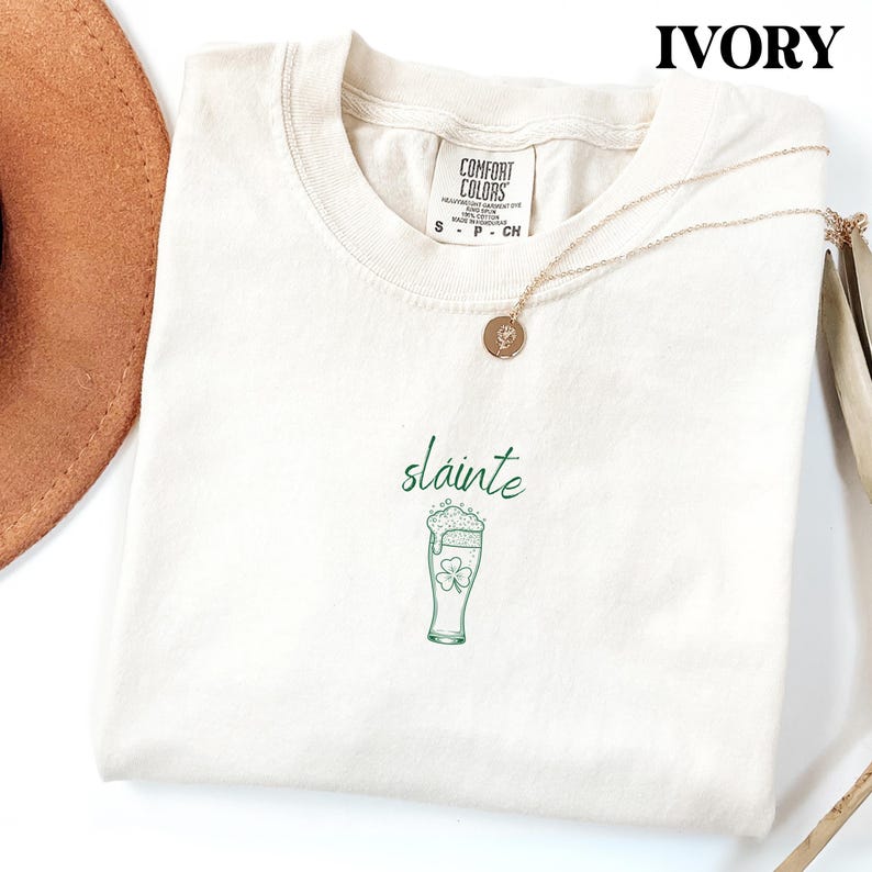 May include: Ivory-colored t-shirt with a crew neck. The shirt features the word "sl&aacute;inte" in green script above a green beer glass with a shamrock. A gold necklace with a round pendant is draped across the shirt.