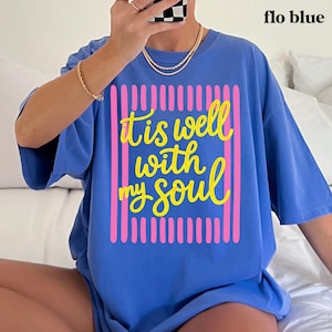 May include: A blue t-shirt with the text "It is well with my soul" in yellow script, framed by pink vertical lines. The shirt is labeled "flo blue" in the upper right corner. The shirt is a casual, oversized style.