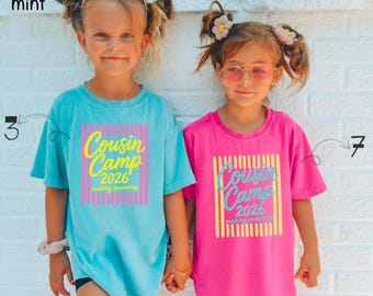 May include: Two short-sleeve t-shirts. The left shirt is chalky mint with a pink striped square and yellow text reading Cousin Camp 2026 making memories. The right shirt is crunchberry pink with a yellow striped square and blue text reading Cousin Camp 2026 making memories.