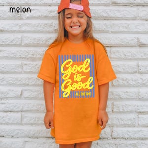 May include: An orange short-sleeved t-shirt featuring a graphic print with vertical blue stripes and the yellow text God is Good ALL THE TIME. The shirt has a crew neckline and a straight hem, providing a simple and classic fit for casual wear.