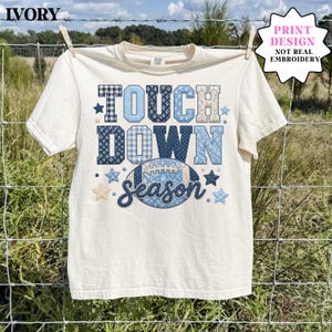 May include: An ivory short-sleeve t-shirt featuring a graphic print with the text "TOUCH DOWN season" in blue and white patterned block letters. The design includes a football graphic and various star shapes, with a note stating it is a print design.