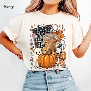May include: An ivory t-shirt featuring a graphic print of a composition notebook, a pencil, an iced coffee in a plastic cup, a maple leaf, a ghost, a pumpkin, and a plaid bow. The design includes small heart and star accents in black and orange.