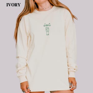 May include: Ivory long-sleeve shirt with the word "sl&aacute;inte" and a green beer graphic. The shirt is a cream color and has a crew neck. The text and graphic are located on the upper chest area.