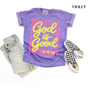 May include: A violet short-sleeve t-shirt with a graphic print that reads God is Good ALL THE TIME in yellow script and block lettering over pink vertical stripes. The shirt is paired with light blue denim shorts and black and white checkered slip-on shoes.