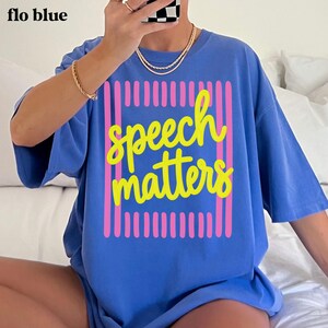 May include: A periwinkle blue short-sleeve t-shirt featuring a graphic design with a rectangular frame made of vertical pink stripes. Centered inside the frame is the text "speech matters" written in a bright yellow, cursive-style font.