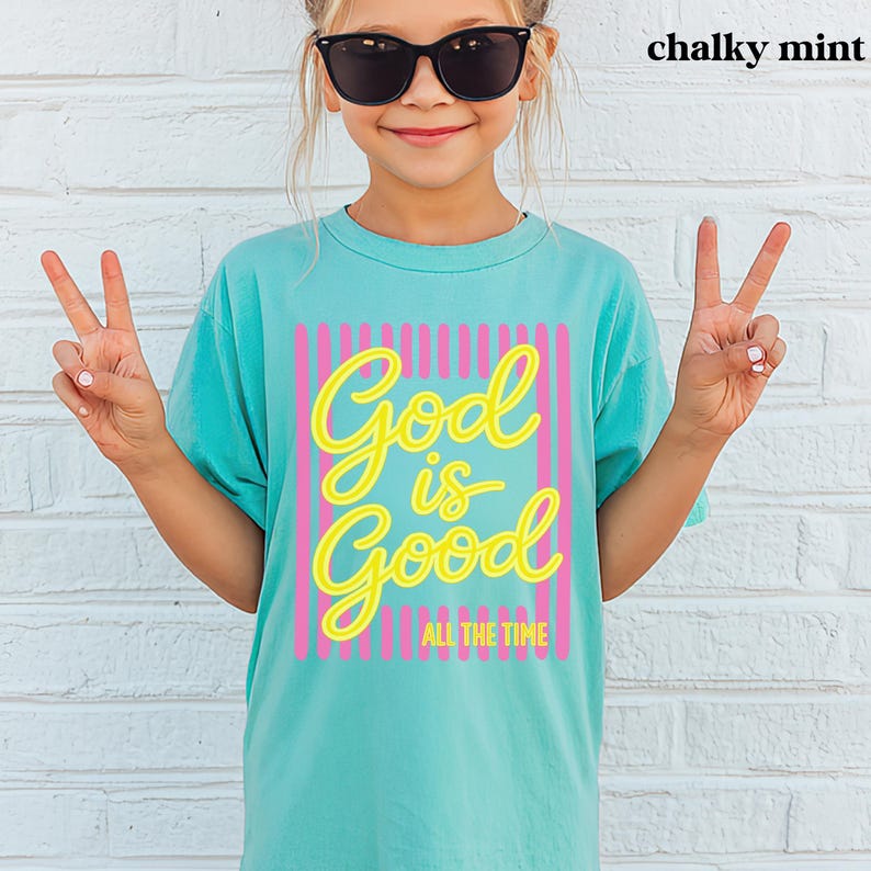 May include: A chalky mint short-sleeve t-shirt featuring a graphic print with vertical pink stripes and the yellow text God is Good ALL THE TIME. The shirt has a crew neckline and a relaxed fit, showcasing a bright and colorful design.