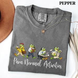 May include: A gray Comfort Colors t-shirt featuring a cartoon illustration of four frogs in various work-related scenarios. The phrase "Para Normal Activities" is printed in white script below the frogs. A silver necklace with a pendant is draped across the shirt.